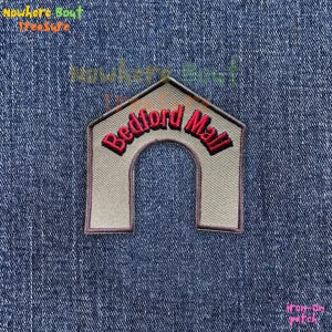 May include: An iron-on patch featuring a beige archway design with the words "Bedford Mall" in red script. The patch is on a blue denim background. The patch is a vintage style.