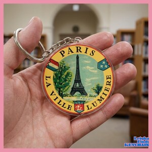 May include: A round keychain featuring a vintage-style design of the Eiffel Tower in Paris, France. The keychain has a yellow background with the words "Paris" and "La Ville Lumiere" around the edges. The keychain is held in a hand.