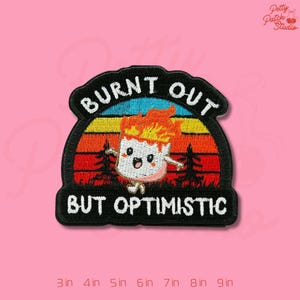 May include: Embroidered patch with the text "BURNT OUT BUT OPTIMISTIC". The design features a cartoon marshmallow on fire against a sunset backdrop. The patch is black with a pink, orange, yellow, and blue sunset.