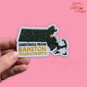 May include: Embroidered patch in the shape of Massachusetts, with the text "GREETINGS FROM BAHSTON MASSACHUSETTS". The patch features a dark green state outline, a yellow star, and white and yellow lettering. The patch is held against a pink background.