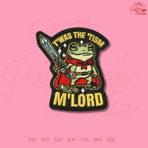 May include: Embroidered patch featuring a green frog in a red cape, holding a sword. The patch reads "T'WAS THE 'TISM M'LORD" in yellow text on a black background. The patch is approximately 3 inches wide.