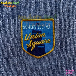 May include: Embroidered patch with a blue shield design. The patch features the text "SOMERVILLE, MA" and "Union Square" in yellow script. A dark blue silhouette of a building is also visible.