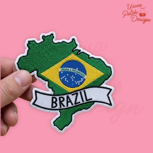 May include: Embroidered patch in the shape of Brazil, featuring the Brazilian flag's colors: green, yellow, blue, and white. The word "BRAZIL" is embroidered on a white banner. The patch is held against a pink background.