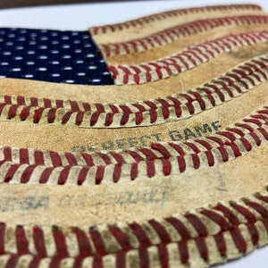 May include: A close-up of a baseball-themed American flag. The flag is made of leather and has red stitching. The words "We Got Game" are visible on the leather.