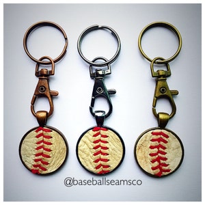 May include: Three keychains with baseball-themed charms. Each charm features a white leather baseball with red stitching. The keychains are attached to different colored metal rings: copper, silver, and gold.