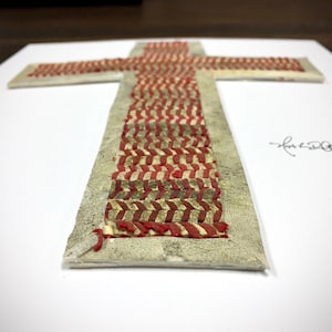 May include: A white cross with a red and gold woven pattern. The cross is mounted on a white background and signed "Lauren Olson".