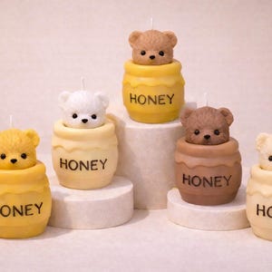 Könnte beinhalten: Eine Sammlung von Bärenkerzen in verschiedenen Farben, darunter Gelb, Braun und Weiß. Jede Kerze zeigt einen Bärenkopf, der aus einem honigtopfförmigen Sockel mit dem Aufdruck "HONEY" herausragt. Die Kerzen sind auf einer weißen Oberfläche angeordnet.