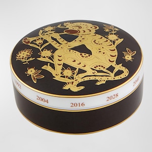 May include: A round, dark brown decorative box with a gold-colored illustration of a tiger and floral elements. The box has a white band with gold text indicating the years 1992, 2004, 2016, 2028, and 2040. The lid is rounded.