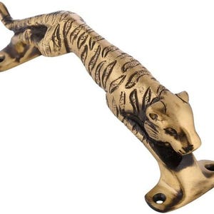 Può includere: Una maniglia per porta color ottone a forma di tigre. La maniglia presenta un design dettagliato di tigre con strisce e una finitura strutturata. La tigre è in posizione di salto, con la testa e la coda sollevate.
