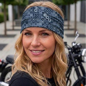Op de afbeelding: Een blauw en grijs patroon hoofdband wordt gedragen door een persoon met blond haar. De hoofdband heeft een herhalend cirkelvormig ontwerp. Een zwarte motorfiets is zichtbaar op de achtergrond.