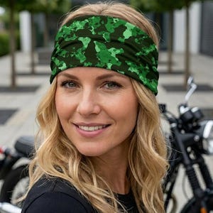Op de afbeelding: Een groene camouflage hoofdband. De hoofdband wordt gedragen door een persoon met blond haar. De achtergrond omvat een motorfiets en bomen. De hoofdband is gemaakt van een zacht, comfortabel materiaal.