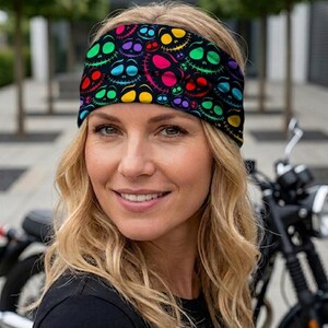 Op de afbeelding: Zwarte hoofdband met een herhaald patroon van lachende schedelgezichten in verschillende kleuren. De schedels zijn groen, geel, rood, paars en roze. De hoofdband wordt gedragen door een persoon met blond haar.