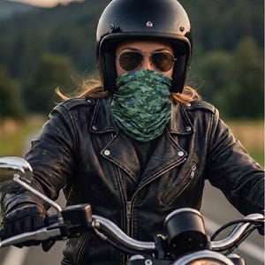 Op de afbeelding: Een persoon met een zwarte motorhelm en zonnebril, met een groen-blauwe camouflage gezichtsbedekking, rijdt op een motor. De persoon draagt een zwarte leren jas. De afbeelding heeft de tekst "Öko-Tex".