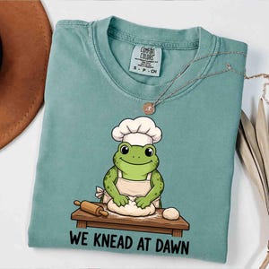 Könnte beinhalten: Ein salbeigrünes T-Shirt mit einer Grafik eines grünen Frosches, der eine weiße Kochmütze und eine Schürze trägt und Teig auf einem Holztisch knetet. Der Text "WE KNEAD AT DAWN" ist in Schwarz unter dem Tisch gedruckt. Mit Comfort Colors Etikett.