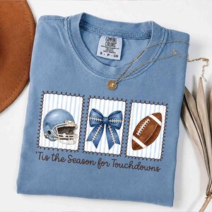 Puede incluir: Una camiseta azul de manga corta con un gráfico de un casco de fútbol americano, un lazo azul y un balón de fútbol dentro de tres marcos rectangulares. El texto 'Tis the Season for Touchdowns' aparece impreso en color marrón.