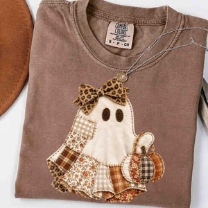 May include: A brown short-sleeve t-shirt featuring a ghost applique with a leopard print bow, a patchwork skirt made of various brown and cream patterned fabrics, and a small pumpkin. The shirt has a Comfort Colors label with size S-P-CH.