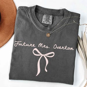 May include: Dark grey t-shirt featuring the text "Future Mrs. Overton" in a cursive font and a pink bow graphic. A silver necklace with a pendant is draped across the shirt. The shirt is folded on a white surface.