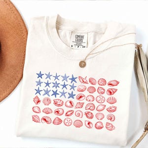May include: Cream-coloured t-shirt with a graphic of the American flag made of blue starfish and red seashells. The shirt has a crew neck and short sleeves. A gold necklace with a pendant is draped on the shirt.