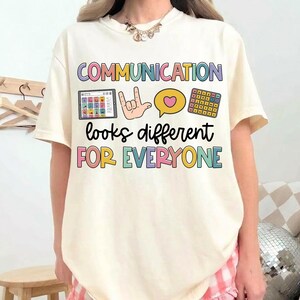 Puede incluir: Una camiseta de manga corta color crema con el texto "COMMUNICATION looks different FOR EVERYONE" en letras coloridas. El gráfico incluye una tableta con iconos, una mano haciendo el signo de te quiero, un bocadillo de corazón y una cuadrícula.