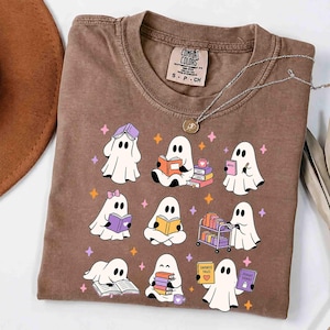 Könnte beinhalten: Ein braunes T-Shirt mit einem Grafikdruck von neun weißen Geistern, die Bücher lesen. Die Geister sind in verschiedenen Posen mit bunten Büchern, einem Bücherwagen und kleinen verstreuten Sternen dargestellt. Das Shirt hat ein Comfort Colors Etikett.