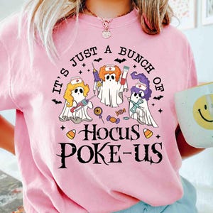 May include: A light pink t-shirt featuring a graphic of three ghosts dressed as nurses with syringes and test tubes. The text reads, "IT'S JUST A BUNCH OF HOCUS POKE-US," surrounded by small bats, stars, and candy corn illustrations in black, orange, and purple.