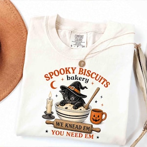 May include: A cream-coloured t-shirt featuring a graphic of a black cat wearing a witch hat in a mixing bowl. The text reads SPOOKY BISCUITS bakery and WE KNEAD EM YOU NEED EM. A candle, a wooden rolling pin, and a jack-o'-lantern mug are included.