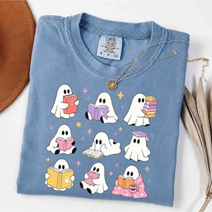May include: A blue short-sleeved t-shirt featuring a graphic print of nine white ghosts reading various colorful books. The ghosts are surrounded by small yellow, pink, and purple stars. The shirt has a Comfort Colors brand label at the neck.