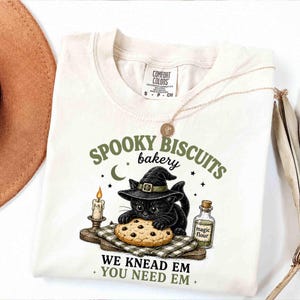 May include: A cream-coloured t-shirt featuring a graphic of a black cat wearing a witch hat, kneading a chocolate chip cookie. The text reads SPOOKY BISCUITS bakery WE KNEAD EM YOU NEED EM, accompanied by a candle and a bottle labelled magic flour.