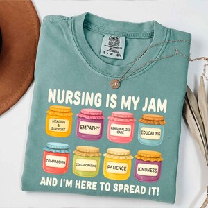 Może przedstawiać: Niebiesko-zielona koszulka Comfort Colors z napisem "NURSING IS MY JAM" i ilustracjami słoików z dżemem. Każdy słoik ma inną etykietę, w tym "Współczucie". Na koszulce leży naszyjnik z wisiorkiem.