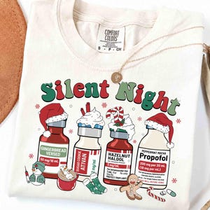 May include: A cream-colored t-shirt featuring a graphic with the text Silent Night and four medical vials labeled Gingerbread Versed, Sugar Cookie Ativan, Hazelnut Haldol, and Peppermint Mocha Propofol, each topped with holiday-themed decorations.