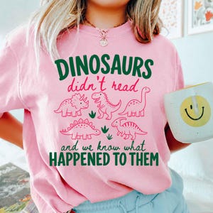 May include: Pink crew neck t-shirt with a green and pink dinosaur graphic. The graphic includes the text "DINOSAURS didn't read and we know what HAPPENED TO THEM" and illustrations of various dinosaurs.