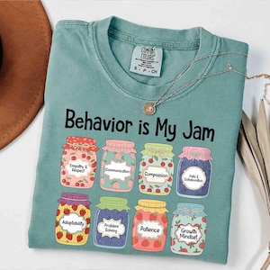 Puede incluir: Una camiseta verde agua con la frase "Behavior is My Jam" en negro. Debajo, ilustraciones de ocho tarros llenos de fruta y etiquetados con atributos positivos como "Empatía y Respeto" y "Mentalidad de Crecimiento". Un collar plateado descansa sobre la camiseta.