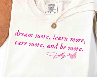 May include: A cream-colored short-sleeve t-shirt with a crew neckline. The shirt features the pink text "dream more, learn more, care more, and be more." followed by the signature "Dolly" and a small butterfly graphic. A gold necklace rests on the shirt.