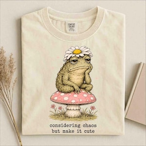 May include: A cream-coloured t-shirt featuring a graphic of a toad wearing a white daisy on its head, sitting on a pink mushroom with white spots. Below the mushroom, the text reads: considering chaos but make it cute. The shirt has a crew neck.