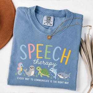 Könnte beinhalten: Ein blaues T-Shirt mit den Worten "SPEECH therapy" in bunten Buchstaben und Meereskreaturen-Grafiken. Das Shirt hat auch den Text "EVERY WAY TO COMMUNICATE IS THE RIGHT WAY". Eine goldene Halskette liegt auf dem Shirt.
