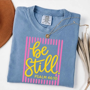 Könnte beinhalten: Ein blaues Comfort Colors T-Shirt mit den Worten "be still" in gelber Schrift, umrahmt von einem rosa Rechteck. Der Text "Psalm 46:10" steht darunter. Eine goldene Halskette mit Anhänger liegt auf dem Shirt.