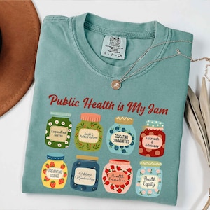 Könnte beinhalten: Ein teal Comfort Colors T-Shirt mit dem Schriftzug "Public Health is My Jam" in roter Schrift. Das Shirt zeigt Illustrationen von Gläsern mit verschiedenen Themen der öffentlichen Gesundheit, darunter "Expanding Access" und "Health Equity".