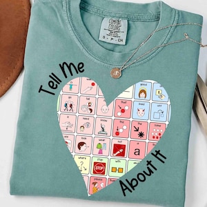 Puede incluir: Una camiseta verde salvia con un gráfico en forma de corazón lleno de una cuadrícula de símbolos e iconos de comunicación. El texto "Tell Me" aparece sobre el corazón y "About It" debajo. Un collar fino de tono dorado descansa en el cuello.