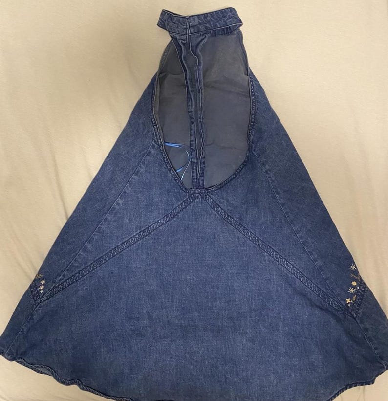 Puede incluir: Un vestido halter de mezclilla azul con falda acampanada. El vestido presenta costuras decorativas a lo largo de las costuras y un peque&ntilde;o bordado floral cerca del dobladillo. El cuello halter est&aacute; abierto por la espalda.