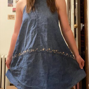Puede incluir: Vestido vaquero sin mangas, cuello halter, en un lavado azul medio. El vestido presenta un sutil bordado floral a lo largo del dobladillo. La silueta en forma de A y el dise&ntilde;o sencillo lo convierten en una prenda vers&aacute;til para uso informal.