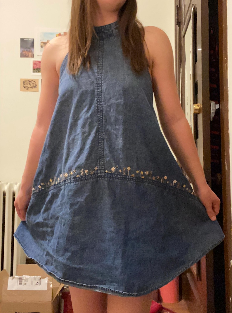 Puede incluir: Vestido vaquero sin mangas, cuello halter, en un lavado azul medio. El vestido presenta un sutil bordado floral a lo largo del dobladillo. La silueta en forma de A y el dise&ntilde;o sencillo lo convierten en una prenda vers&aacute;til para uso informal.