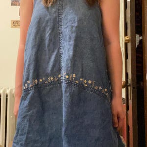 Puede incluir: Vestido vaquero azul con cuello halter y bordado floral en el bajo. El vestido sin mangas tiene un cuello alto y una falda acampanada. El bordado presenta peque&ntilde;as flores estilizadas en tonos dorados y marrones. Parece estar hecho de mezclilla de peso medio.