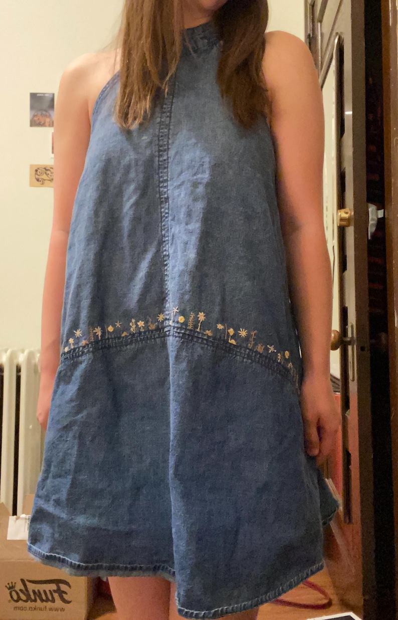 Puede incluir: Vestido vaquero azul con cuello halter y bordado floral en el bajo. El vestido sin mangas tiene un cuello alto y una falda acampanada. El bordado presenta peque&ntilde;as flores estilizadas en tonos dorados y marrones. Parece estar hecho de mezclilla de peso medio.