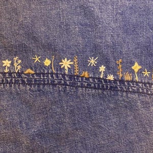 Puede incluir: Primer plano de una tela vaquera azul con un dise&ntilde;o bordado. El bordado presenta una serie de peque&ntilde;as formas florales y estrelladas estilizadas en tonos beige y dorado, cosidas a lo largo de una costura.