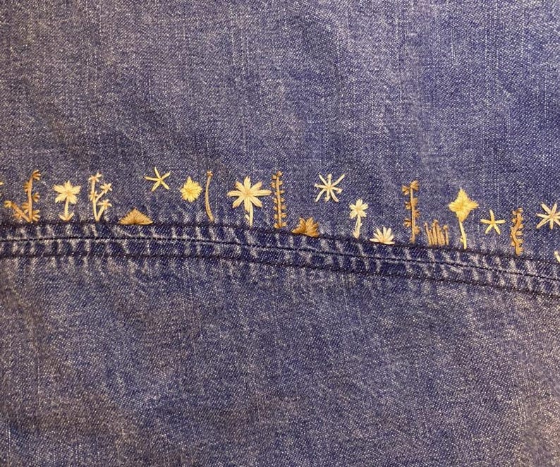 Puede incluir: Primer plano de una tela vaquera azul con un dise&ntilde;o bordado. El bordado presenta una serie de peque&ntilde;as formas florales y estrelladas estilizadas en tonos beige y dorado, cosidas a lo largo de una costura.