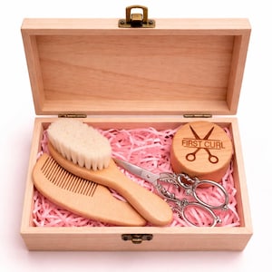 May include: A wooden box containing a baby grooming kit. The kit includes a wooden brush, comb, scissors, and a wooden disc with the text "FIRST CURL". The box is filled with pink paper shreds.