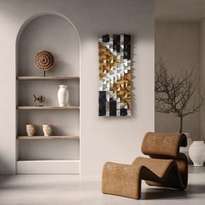 May include: A modern wall art piece with a geometric design. The artwork features a mosaic of small, square wooden blocks in black, gold, and silver. The piece is rectangular and mounted on a neutral-colored wall. A brown leather chair is in the foreground.