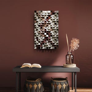 May include: Geometric wall art with a textured, three-dimensional design in shades of white, gray, and brown. The artwork is rectangular and hangs on a maroon wall. A dark gray table below holds an open book, decorative baskets, and a vase with dried flowers.