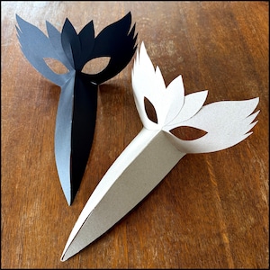 May include: Two pointed masks, one solid black and one solid white, each featuring a sharp, elongated nose section and wing-like eye cutouts. The masks are constructed from folded paper or cardstock, displaying a symmetrical, angular design.