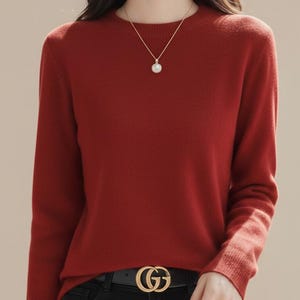 Peut inclure: Un pull rouge brique à manches longues et col rond. Le pull est associé à un jean noir et une ceinture noire avec une boucle dorée. Un collier en or avec un pendentif perle est également visible.